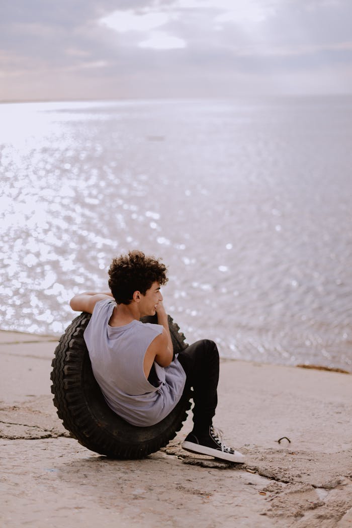gallery-3 Teen sitting on a tire by the seashore, enjoying a peaceful moment with the ocean view.